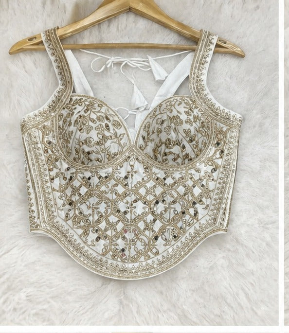 An ornate white and gold embroidered bustier-style top hanging on a wooden hanger against a white faux-fur background.