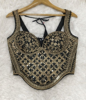 Black and gold heavily embroidered sleeveless crop top blouse with floral patterns, displayed on a wooden hanger against a white faux fur background.