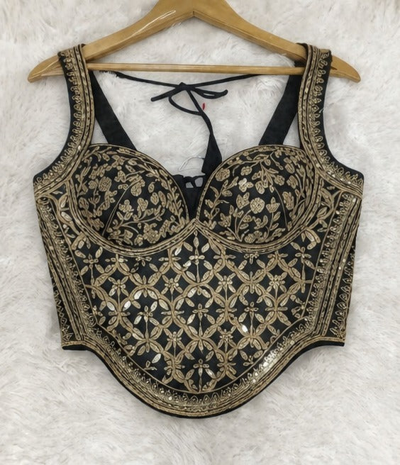 Black and gold heavily embroidered sleeveless crop top blouse with floral patterns, displayed on a wooden hanger against a white faux fur background.