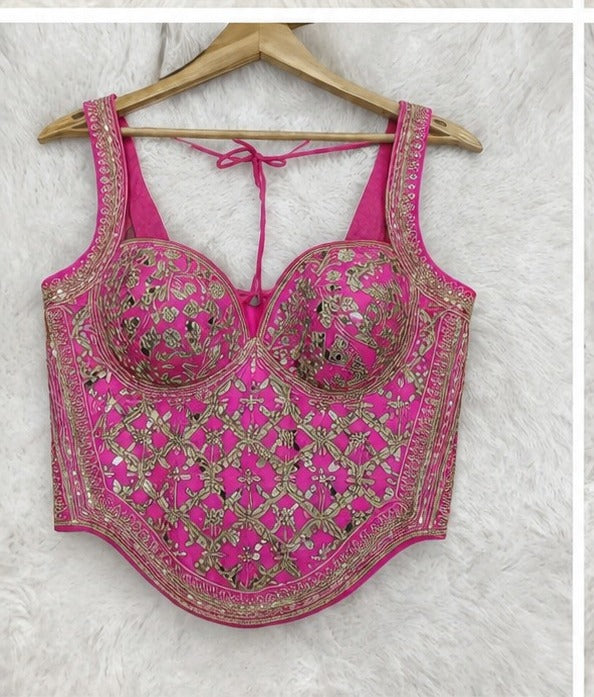 A bright pink sleeveless blouse top with intricate gold and silver mirror work embroidery, displayed on a wooden hanger against a white faux fur background.