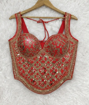 A red sleeveless corset-style top with gold floral embroidery and mirror work, hanging on a wooden hanger against a white faux-fur background.
