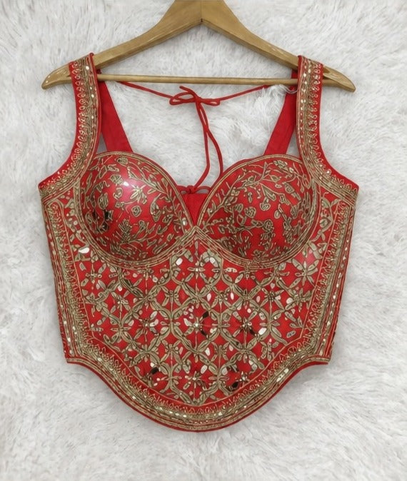A red sleeveless corset-style top with gold floral embroidery and mirror work, hanging on a wooden hanger against a white faux-fur background.
